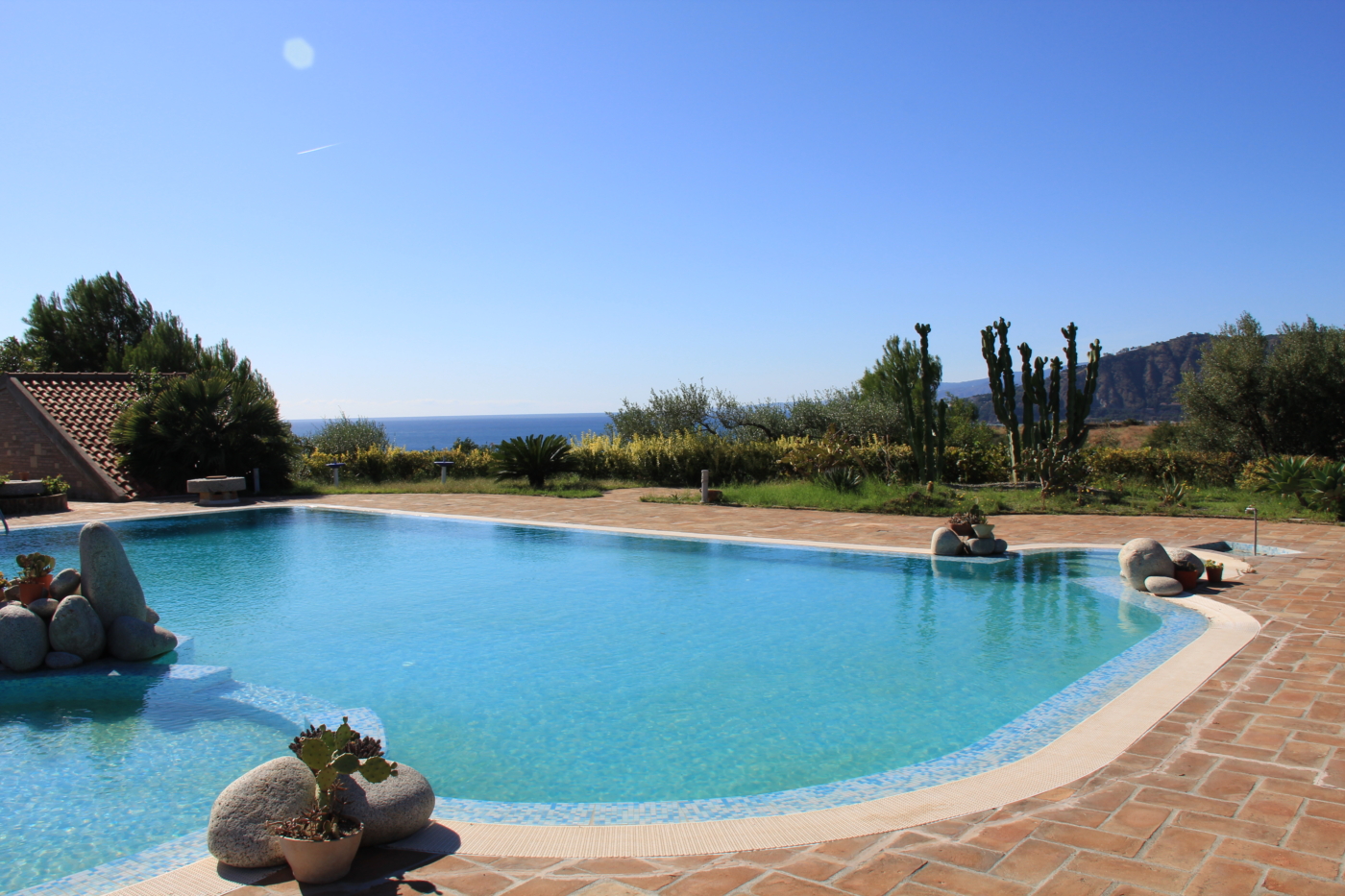 Piscina di Borgo Piazza a mosaico azzurro con bordo in cotto, massi e cactus, uliveti e mare sullo sfondo
